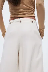 Pantalón sastrero cropped de tiro alto, color beige, con piernas amplias, pliegues frontales, bolsillos al bies y trabillas en la cintura con detalles de botones.