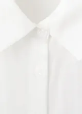 Camisa blanca de manga larga con cuello camisero, cierre frontal de botones y un llamativo detalle de nudo drapeado en el lateral delantero.