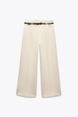 Pantalón culotte color beige de tiro medio, con trabillas y cinturón fino de efecto piel ajustable con hebilla metálica. Presenta detalle de raya planchada y cierre con cremallera oculta en costura.