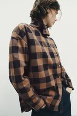 Camisa de manga larga con estampado de cuadros en tonos marrón y negro. Presenta un diseño cruzado con cuello vuelto y cierre frontal de botones, con un corte asimétrico en el bajo.