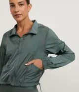 Campera cortavientos deportiva de corte cropped, color verde oscuro. Presenta capucha, cierre frontal con cremallera, mangas largas con puños elásticos y cordón ajustable en el ruedo.