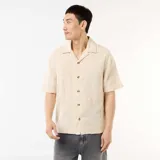 Camisa de manga corta color beige con cuello tipo campamento, cierre frontal de botones y diseño de textura en punto fantasía.