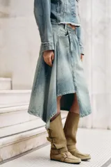 Falda midi de denim con lavado claro, tiro medio y cintura cruzada con cierre asimétrico de cremallera y botones metálicos. Presenta bolsillos delanteros y bajo asimétrico.