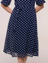 Vestido midi azul marino con estampado de lunares blancos, confeccionado en gasa semitraslúcida. Tiene escote redondo, mangas cortas acampanadas con volantes en los hombros, lazo ajustable en la cintura y falda con vuelo y forro interior.