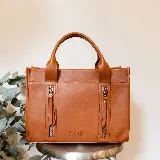 Cartera de cuero color tostado, estructurada, con doble asa de mano, correa desmontable y dos bolsillos frontales verticales con cierre. Presenta detalles de costuras pespunteadas y el logo de la marca grabado en la parte inferior.
