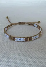 Pulsera tejida artesanalmente con cuentas de colores blanco, marrón, dorado y un detalle en celeste, montada sobre cordón marrón ajustable.