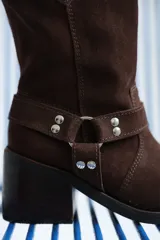 Bota de caña media confeccionada en cuero gamuzado color chocolate. Presenta un diseño de estilo western con taco cuadrado de 7 cm y una correa decorativa en el tobillo con herrajes metálicos plateados.