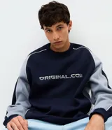 Buzo de corte boxy con cuello redondo y mangas largas raglán. Presenta un diseño bicolor en azul marino y gris, con detalles de piping en contraste y lettering bordado en el pecho.
