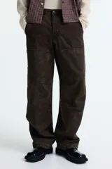 Pantalón estilo carpenter de corte holgado, confeccionado en tejido de algodón color marrón oscuro. Presenta tiro caído, bolsillos laterales de parche, trabilla funcional en una pierna y costuras reforzadas visibles sobre y debajo de las rodillas.