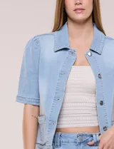 Camperita de denim color celeste con efecto lavado suave, cuello camisero clásico y cierre frontal de botones metálicos. Tiene mangas cortas con fruncido en los hombros, bolsillos delanteros bajos y costuras estructuradas.