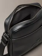 Bandolera reporter Calvin Klein de poliuretano color negro, con correa ajustable al hombro, cierre superior con cremallera, bolsillo frontal externo y logo monograma grabado en la parte inferior.