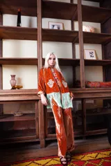 Camisa de seda en color verde petróleo, con corte recto y estructura clásica. Presenta un diseño con apliques bordados en tono borravino y detalles de flecos largos en la parte frontal inferior.