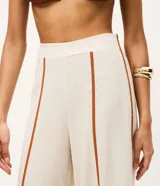 Pantalón largo tipo palazzo de color beige, confeccionado en mezcla de viscosa y lino. Presenta tiro alto, cintura elastizada en la parte posterior y aberturas frontales en las piernas con ribetes en tono marrón contrastante.