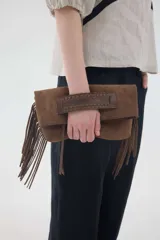 Cartera tipo sobre de gamuza color marrón, con solapa plegable, flecos colgantes en los laterales y una tira de cuero rígida en la parte frontal para sujetar con la mano.