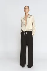 Camisa de cuello solapa y manga larga con puños anchos, confeccionada en tejido satinado color beige. Presenta cierre frontal con botones ocultos y bajo ajustable mediante cordones del mismo tejido.