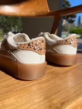 Zapatillas urbanas de cuero color beige y blanco, con tachas plateadas en los laterales y suela de goma color caramelo. Diseñadas en Uruguay y producidas en Brasil.