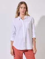 Camisa blanca de corte clásico con textura de líneas verticales, cuello con solapa, cierre frontal con botones y bolsillo en el pecho. Presenta mangas tres cuartos regulables con presilla y botón.