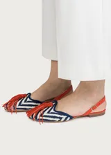 Sandalias planas destalonadas confeccionadas en rafia trenzada con patrón de zigzag en tonos azul marino y crudo. Tienen punta cerrada con una borla decorativa de flecos naranjas en el empeine, y una tira trasera ajustable de cuero naranja con hebilla metálica dorada.