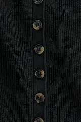 Chaleco negro de punto acanalado, sin mangas, con cuello redondo y cierre frontal de botones. Presenta un diseño entallado con bajo en peplum.