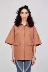 Sobrecamisa unisex color marrón claro, de manga corta y corte ancho. Presenta abertura frontal con botones tono sobre tono, dos bolsillos de parche en la parte inferior delantera y un bolsillo de ribete en el pecho. Logotipo de la marca estampado en contraste en el pecho y estampado grande en dos tonos en la espalda.