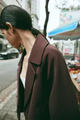 Trench corto de color marrón oscuro, con cuello de solapa, cierre frontal cruzado con botones y mangas tres cuartos con detalle de vuelta, trabilla y botón. Cuenta con bolsillos delanteros de vivo.