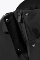 Anorak corto de color negro con acolchado interior, cuello subido y capucha. Presenta mangas largas con puños elásticos, bolsillos delanteros con cierre y bajo ajustable con cordones. Cierre frontal con cremallera termosellada y botones a presión.