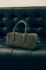 Cartera tipo satchel de diseño rectangular, confeccionada en gamuza color verde oliva. Cuenta con doble asa corta de mano, cierre superior con cremallera metálica y logo de la marca grabado en el frente.