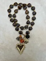 Collar de cuentas de resina en forma de corazón en tonos marrones y ámbar, intercaladas con pequeñas cuentas doradas. Presenta un conjunto de dijes colgantes que incluyen un corazón grande de metal dorado con textura martillada, un dije de corazón sagrado, un corazón pequeño esmaltado y cuentas adicionales en tonos ámbar y negro.