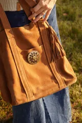 Cartera tote de gamuza color marrón claro, con bolsillo exterior con broche metálico y hebilla decorativa, y detalle de flecos colgando de un lateral.