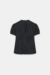 Camisa negra con estampado de lunares blancos, cuello redondo con escote en pico, mangas cortas abullonadas y detalle de volantes frontales. Cierre frontal con botones.