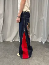 Pantalón de jean azul raw con diseño de corte versátil, ajustable mediante cierres laterales que revelan un panel interior rojo. Presenta una estampa gráfica de gran tamaño en la parte trasera con detalles bordados, costuras reforzadas y etiqueta de cuero en la cintura.