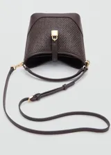Bolso tipo bucket de tamaño pequeño, confeccionado con tejido de rafia color marrón con efecto texturizado y detalles en cuero sintético liso del mismo tono. Cuenta con un asa corta superior, herrajes metálicos dorados, cierre de imán y un detalle decorativo con candado dorado en la parte frontal.