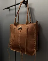 Cartera tipo tote de cuero color tostado, con diseño artesanal que incluye detalles de trenzado en los bordes y en el centro, y un cierre decorativo con un aplique en forma de cuerno.