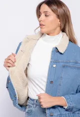 Campera de jean celeste con cuello y forro de corderito color crema, corte cuadrado, cierre frontal con botones metálicos, dos bolsillos de parche en el pecho y elástico en el ruedo.