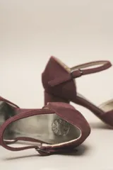 Zapato de tacón color bordó, confeccionado en terciopelo suave, con diseño de pulsera en el tobillo y cierre de hebilla. La punta es cerrada y presenta detalles de líneas cosidas. El tacón es ancho y forrado, con una altura de 7 cm.