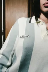 Chomba de manga larga con diseño de rayas verticales en color blanco y verde seco. Presenta cuello tipo polo con cierre de botones y parches decorativos en el pecho.