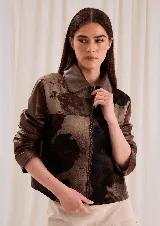 Chaqueta corta con diseño patchwork en tonos chocolate, negro y crudo, confeccionada en cuero combinado con pelo vacuno. Cuenta con cuello clásico, mangas largas y cierre metálico frontal.