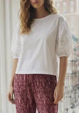 Remera blanca de algodón con mangas cortas abullonadas que presentan detalles de encaje calado y bordado inglés.