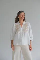Blusa blanca de corte holgado con cuello mao y cierre frontal de botones. Presenta mangas largas abullonadas con puños elásticos y un delicado bordado en color beige en la parte superior delantera.