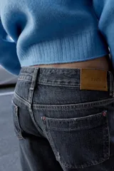 Jeans de mezclilla azul oscuro con efecto desgastado, tiro medio, corte relajado y pernera ancha. Presentan un bajo ancho doblado hacia arriba, revelando el revés del tejido de color beige claro con un detalle de costura amarilla.