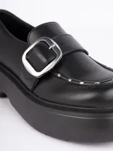 Zapatos tipo mocasines negros con plataforma de 6cm, hebilla metálica plateada y pequeños detalles plateados en la capellada.