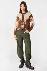 Cardigan de punto color crudo con detalles en marrón oscuro. Presenta cuello redondo, cierre frontal con botones tipo alamares y puños y ruedo acanalados con rayas horizontales. Incluye un pequeño aplique bordado en la manga.