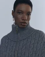 Chaleco largo de punto trenzado color gris con cuello alto y cierre frontal con cremallera metálica.