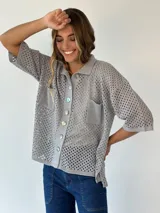 Camisa de punto calado con lúrex, de diseño holgado y manga corta. Cuenta con cuello tipo polo, cierre frontal con botones nacarados y dos bolsillos aplicados en el pecho. Su tejido abierto le otorga una apariencia ligera y brillante ideal para el verano.