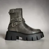 Borcego de cuero sintético en color gris con acabado desgastado, presenta tres correas con hebillas metálicas ajustables y una suela track de goma negra de gran volumen.