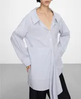 Camisa de algodón a rayas blancas y azules con diseño desestructurado, cuello italiano, manga larga y cierre de botones. Cuenta con una pieza asimétrica multiposición en la parte frontal.