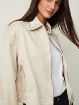 Campera cruda de gabardina con cierre frontal, bolsillos con cierre y cuello mao adornado con tachas plateadas.