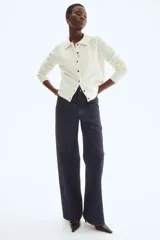 Cárdigan corto de punto suave color crema, con cuello camisero, abotonadura frontal con botones marrones y remate acanalado en puños y bajo.