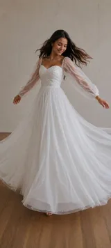 Vestido largo de tul blanco con falda amplia y fluida. Presenta un corsé con escote corazón, drapeado y decorado con apliques de perlas. Incluye mangas largas abullonadas de tul transparente con detalles de perlas.
