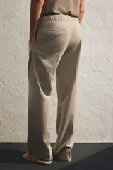 Pantalón de corte relajado color beige claro, con cintura elástica ajustable con cordón oculto y pliegues frontales. Presenta bolsillos laterales y bolsillos traseros ribeteados.